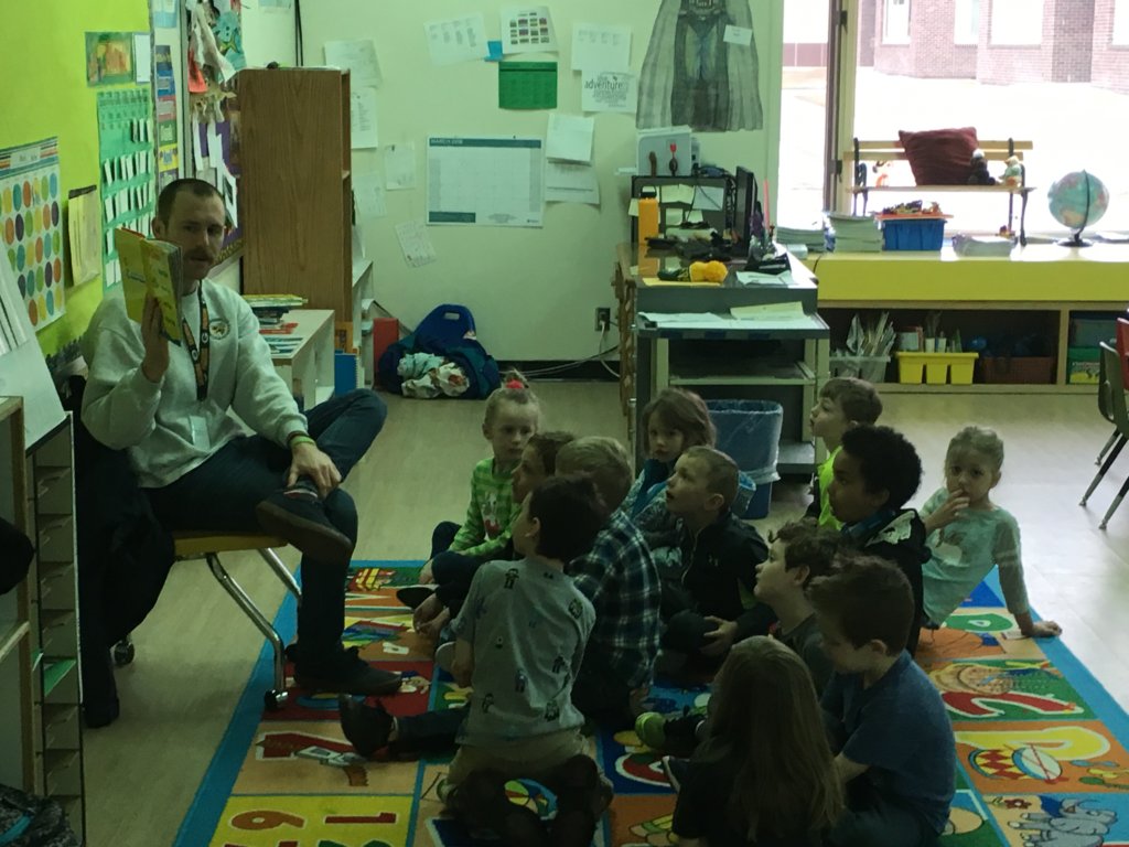 Mr. Harcharik sits in a chair and reads some Dr. Seuss to his Kindergarten class who are all seated on a carpet