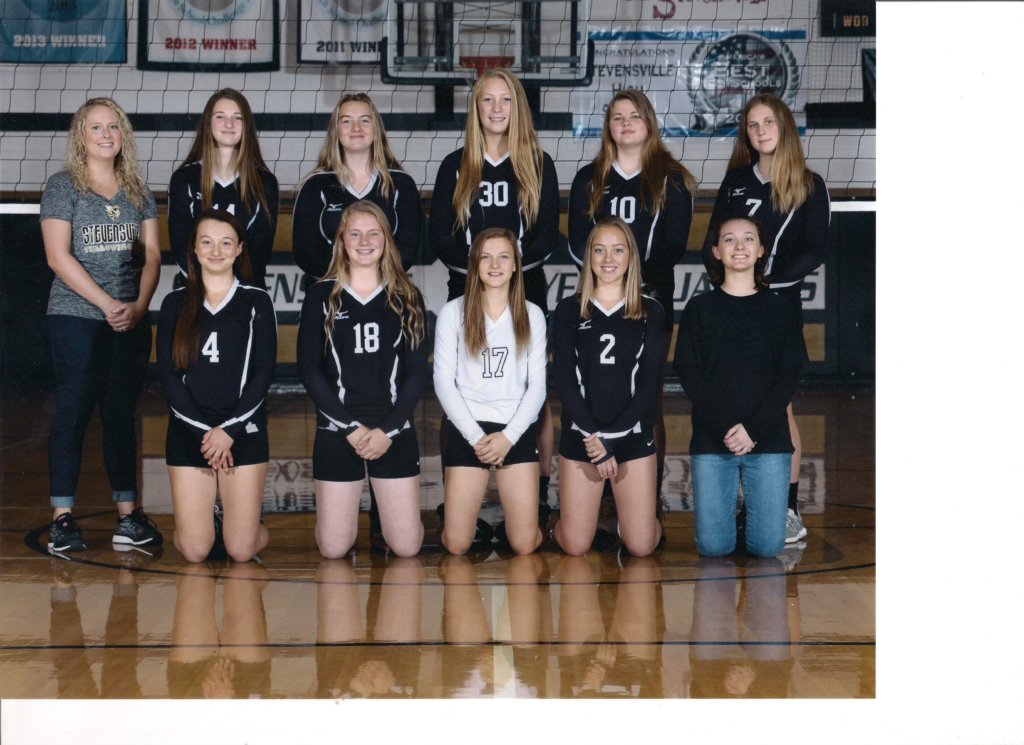 The 2019 Froshmore Team in black jerseys in front of a volleyball net with coaches standing on each side of the team