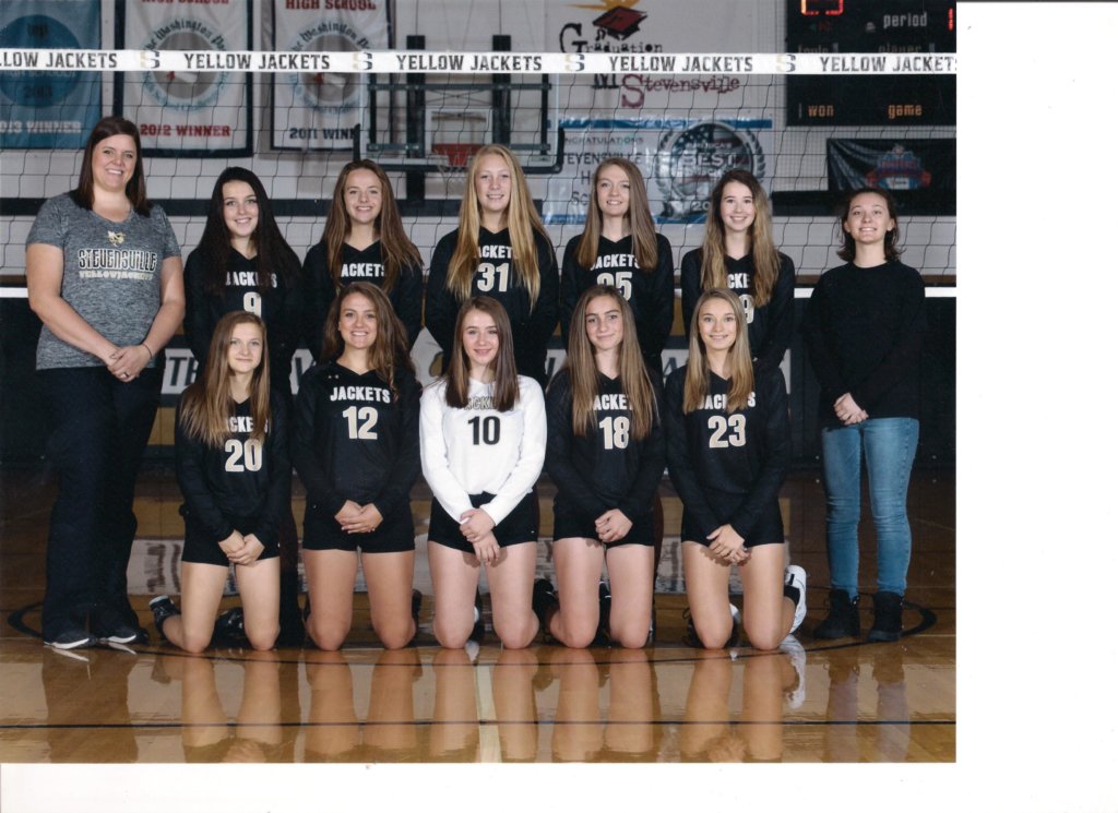 The 2019 Junior Varsity Team in black jerseys in front of a volleyball net with coaches standing on each side of the team