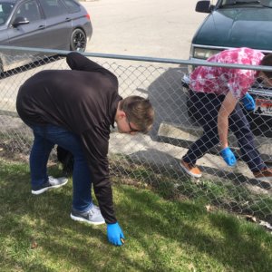 Two students clean up up cigarette buts from the ground near a fence line