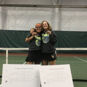 Jackie and Erin celebrate their medals standing behind the brackets showing the entire tournament with a tennis court behind them